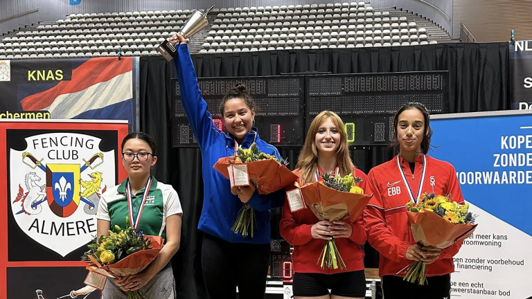 Vier jonge vrouwen poseren met bloemen en medailles; de tweede van links houdt een beker omhoog.