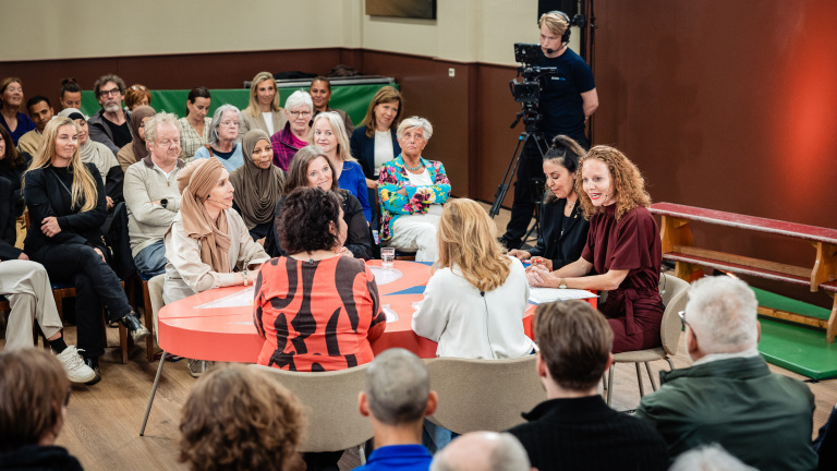 Een groep mensen zit rond een tafel in een discussiepanel, omringd door publiek, met een cameraman op de achtergrond.