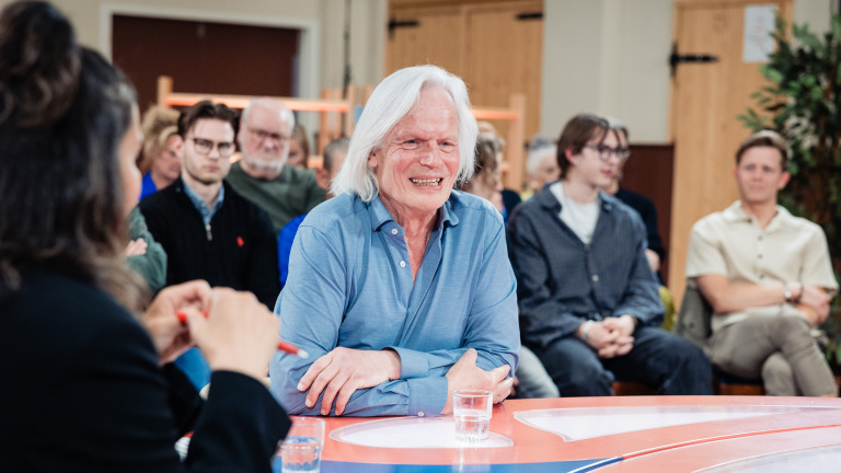 Een oudere man met wit haar en een blauwe overhemd glimlacht tijdens een discussie aan tafel, omringd door publiek.