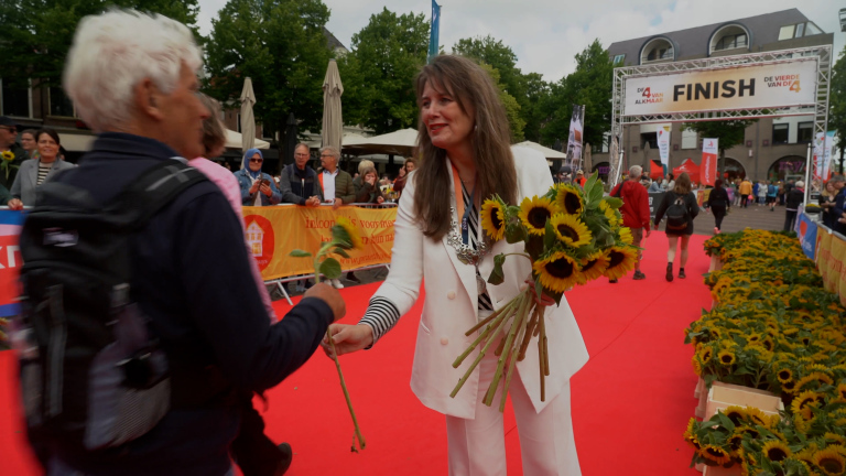 Een vrouw in een witte outfit geeft zonnebloemen aan een deelnemer bij de finish van een evenement met een rode loper.