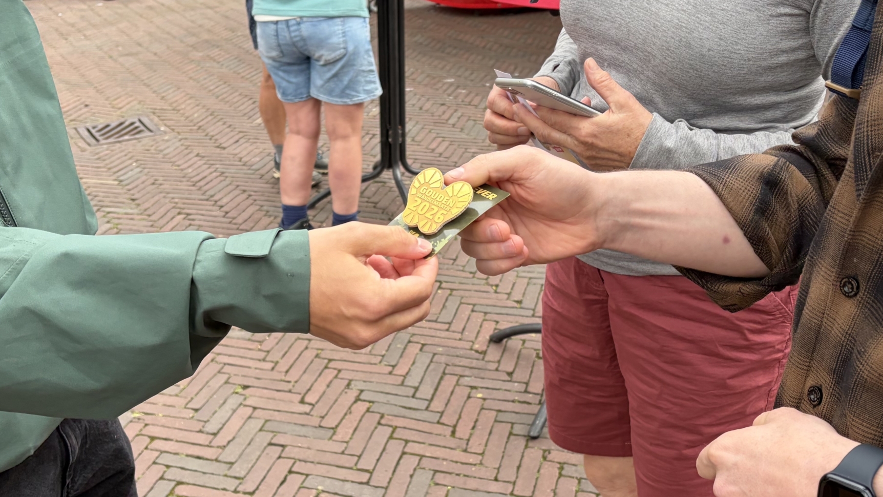 Twee mensen wisselen een gouden medaillekaartje uit op een plein, terwijl anderen op de achtergrond staan.