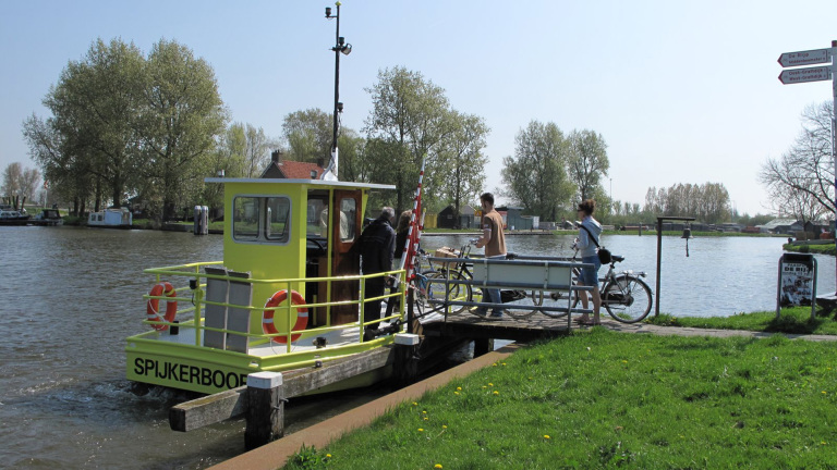 Geel veerbootje genaamd Spijkerboor vervoert personen en fietsen over een rivier bij zonovergoten groen landschap.