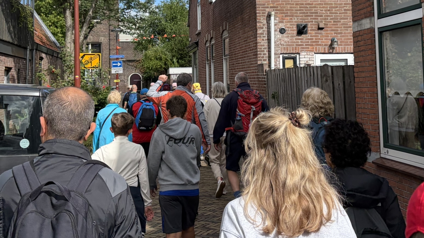 Een groep mensen loopt door een smalle straat tussen bakstenen huizen onder een blauwe lucht met wolken.