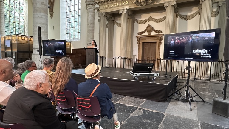 Presentatie in een historische zaal met een vrouw die spreekt vanaf een podium, omringd door een publiek.