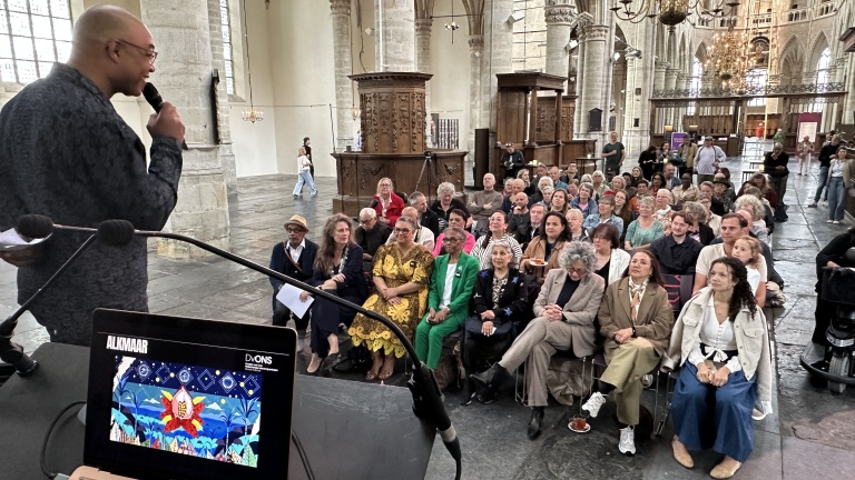 Een spreker spreekt in een kerk voor een aandachtig publiek. Op de voorgrond is een laptop zichtbaar met een kleurrijke afbeelding.
