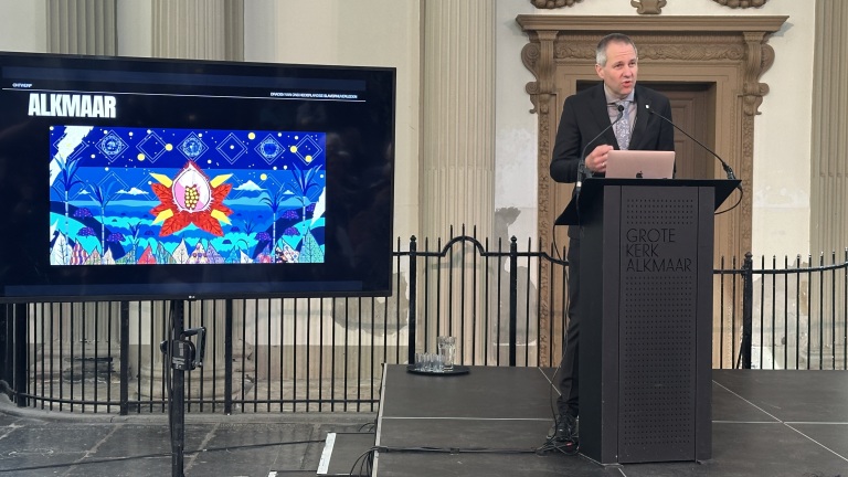 Man spreekt bij een podium in Grote Kerk Alkmaar; scherm toont kleurrijk kunstwerk met opschrift "ALKMAAR".