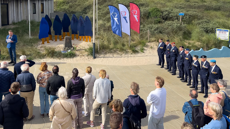 Mensen nemen deel aan een ceremonie buiten bij een monument, met verschillende vlaggen en uniformen.