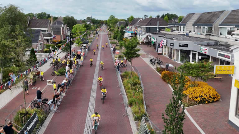Wielerwedstrijd in dorpsstraat, renners in gele shirts fietsen langs afzettingen en toeschouwers, met winkels rechts