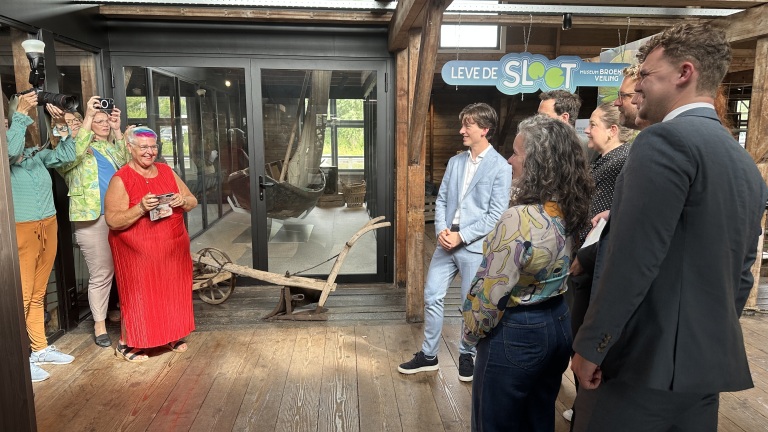 Groep mensen poseert lachend voor enkele fotografen in een houten gebouw met een bord met de tekst "Leve de Sloot".