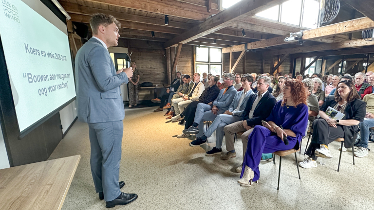 Man in een blauw pak geeft een presentatie aan een aandachtig publiek in een houten zaal, met tekst op een scherm.