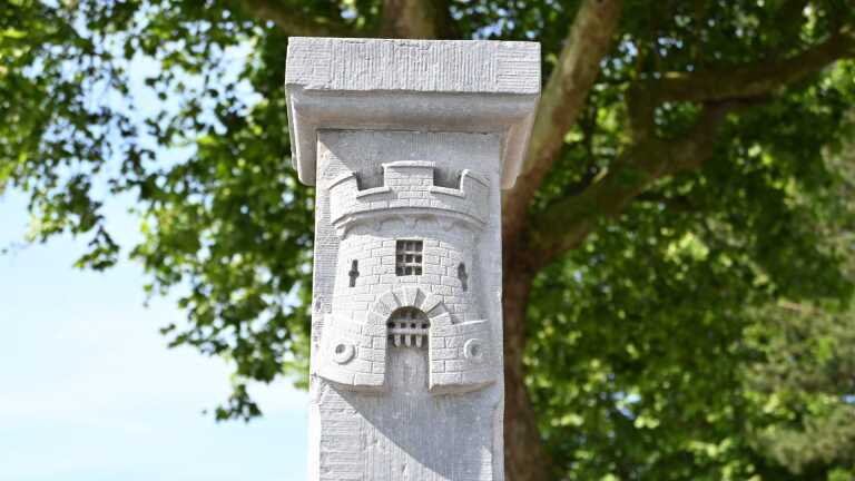 Stenen paal met gebeeldhouwde toren, omringd door groene boombladeren tegen een blauwe hemel.