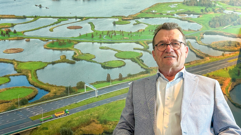 Man met bril en lichtblauw colbert staat voor een muur met een grote foto van een weg en een waterrijk landschap.