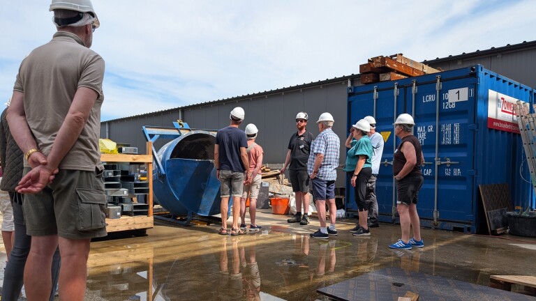 Groep mensen met helmen staat bij een bouwplaats, luisterend naar een gids bij een blauwe container.
