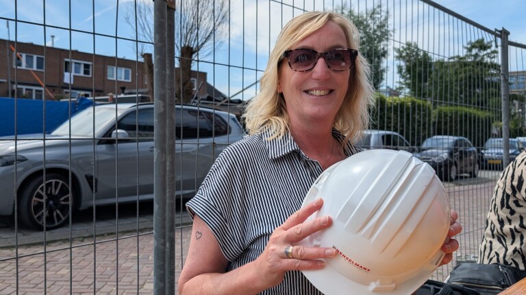 Vrouw met zonnebril en gestreept overhemd houdt een witte bouwhelm vast bij een bouwplaats.
