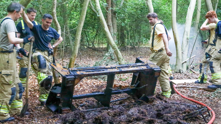 Brandweerlieden inspecteren uitgebrande apparatuur in een bos, met rook op de achtergrond.