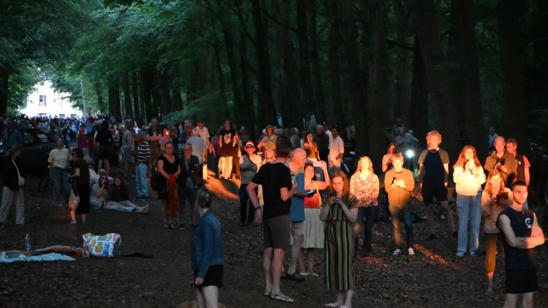 Mensen staan en zitten in een bos, deels verlicht door kunstlicht, tijdens een avondbijeenkomst.