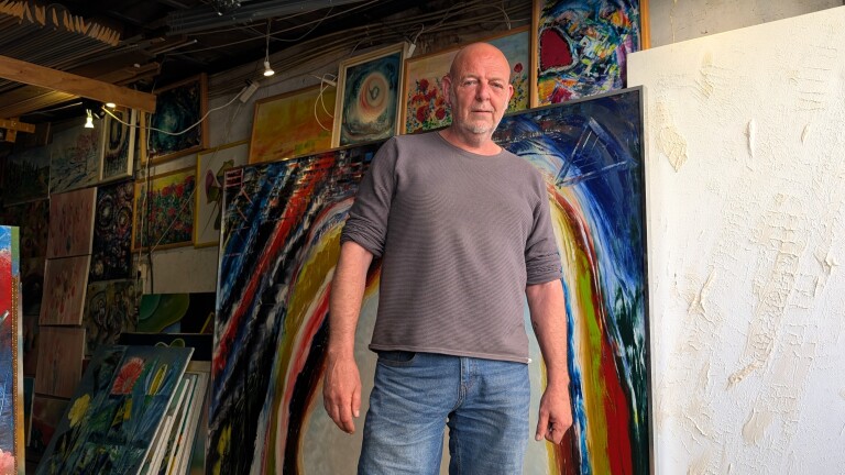 Man in een grijze trui staat voor een kleurrijk schilderij in een kunststudio vol met kunstwerken.