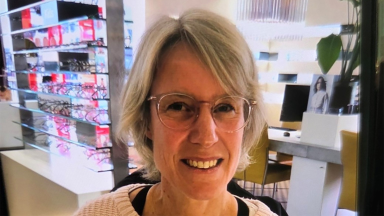 Vrouw met bril glimlacht in een brillenwinkel, omgeven door displays met monturen.