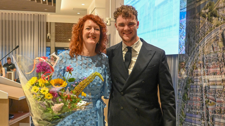 Een vrouw in een blauwe jurk met een boeket bloemen en een man in een donker pak staan samen te glimlachen.