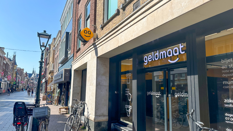 Geldmaat-filiaal aan een zonnige winkelstraat met enkele fietsen geparkeerd voor het gebouw.