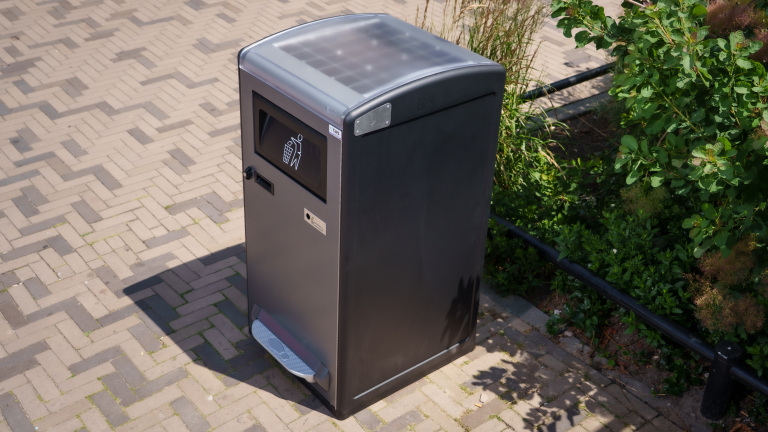 Een moderne vuilnisbak met zonnepanelen staat op een betegelde stoep naast groene planten.
