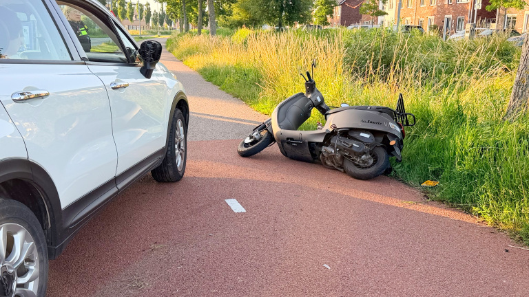 Een witte auto met een beschadigde zijspiegel naast een gevallen scooter op een rood fietspad, met gras en huizen op de achtergrond.