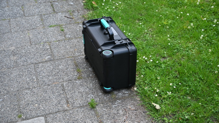 Zwarte koffer met blauwe handgreep en wieltjes staat op een stoep naast gras met witte bloemetjes.