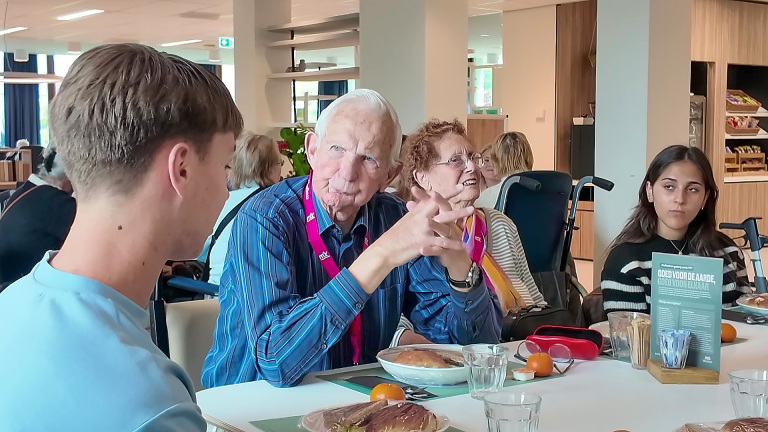 Mensen van verschillende generaties zitten samen aan een tafel in een eetzaal, in gesprek met elkaar.