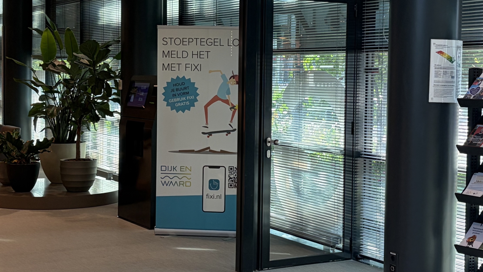 Ganggebied met planten, een informatiebord over Fixi, folderrekken en stoelen naast grote ramen met jaloezieën.