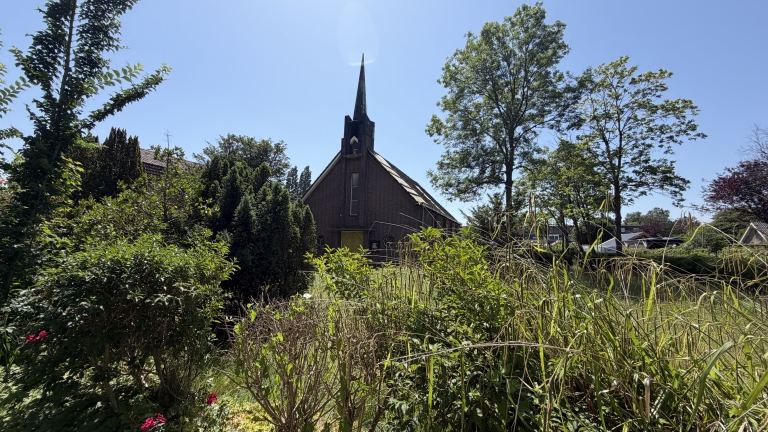 Een kleine bakstenen kerk met hoge torenspits, omgeven door groene bomen en struiken op een zonnige dag.