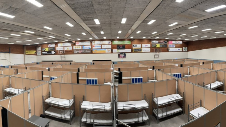 Grote ruimte met rijen stapelbedden gescheiden door kamerschermen, waarschijnlijk een tijdelijke opvanglocatie.
