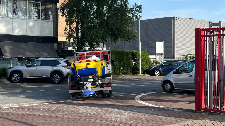 Een voertuig met een geel-blauwe strooimachine rijdt over een straat met geparkeerde auto's en gebouwen op de achtergrond.
