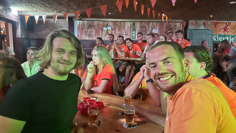 Mensen in oranje shirts zitten in een café met vlaggetjes aan het plafond, waar ze genieten van het kijken naar een wedstrijd.