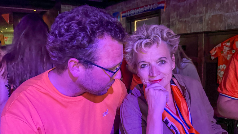 Twee mensen in een kleurrijke ruimte, met oranje kleding die een feestelijke sfeer suggereert.