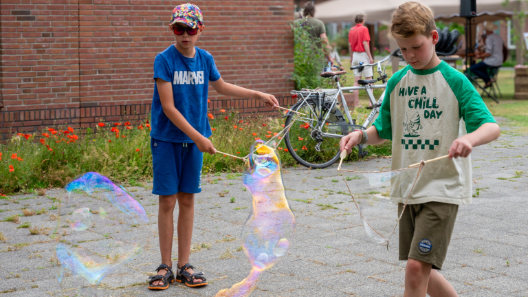 Twee jongens maken zeepbellen met stokken op een plein, met fietsen en een bakstenen muur op de achtergrond.