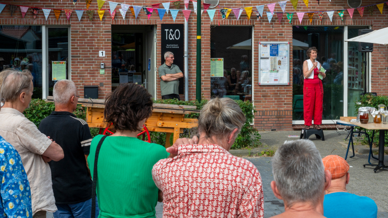 Een vrouw spreekt voor een publiek bij een buurtcentrum, met kleurrijke vlaggetjes en tafels met potten in de omgeving.
