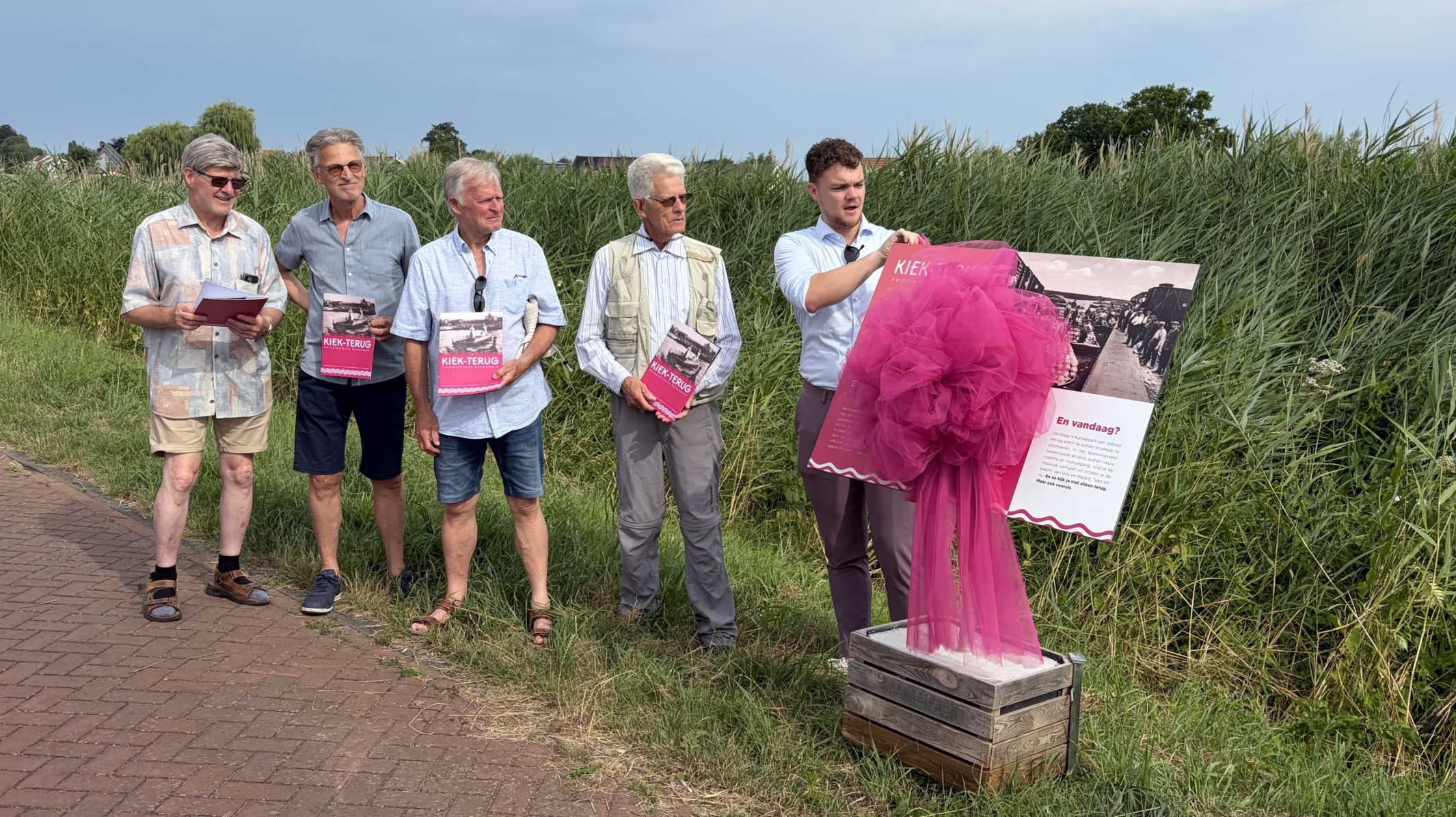 Vijf mannen staan naast een paneel met een roze doek; drie houden een brochure vast. Ze bevinden zich op een pad bij hoog gras.