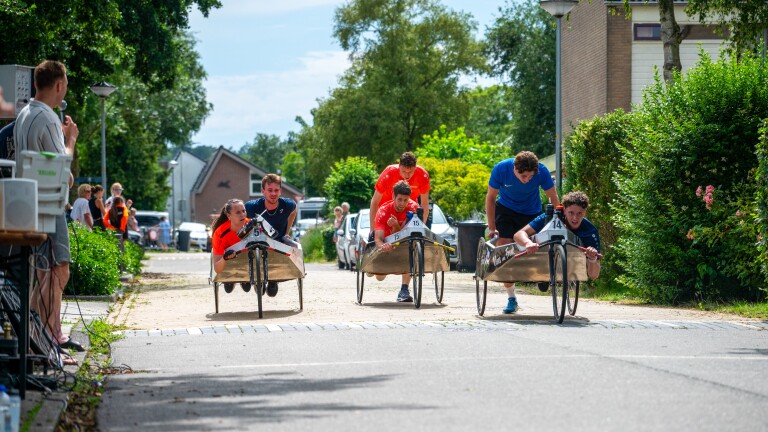 Mensen racen in zelfgemaakte karren over een straat tijdens een evenement met toeschouwers langs de kant.