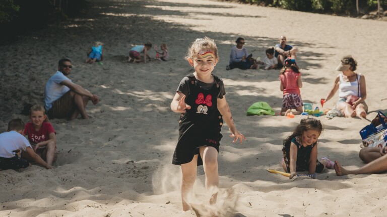Een meisje met geschminkt gezicht rent op een zanderig strand, omringd door spelende kinderen en volwassenen.