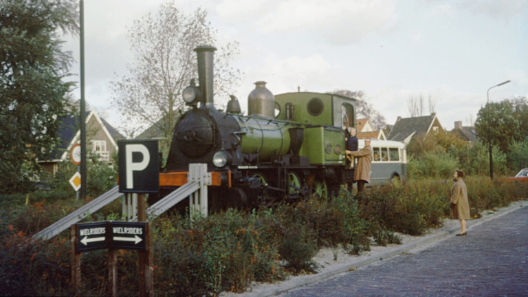 Een groene stoomlocomotief staat op een verhoogd spoor met naastgelegen borden en omringd door struiken; een ouder en kind staan erop, terwijl een vrouw ervoor kijkt.