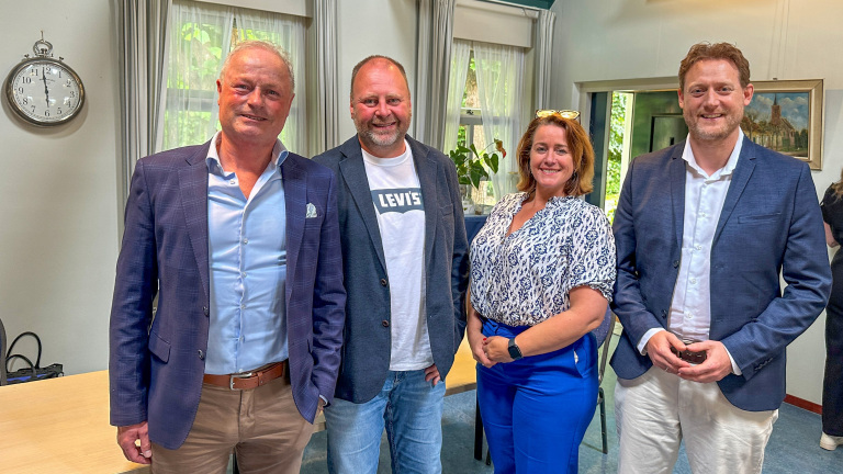 Vier mensen poseren binnen voor een foto, gekleed in semi-formele kleding, met een klok aan de muur op de achtergrond.
