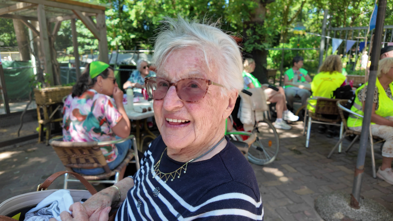 Oudere vrouw met witte haren en bril glimlacht, zittend buiten met andere mensen aan tafels in een zonnige tuin.