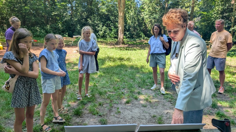 Groep mensen, waaronder kinderen, verzameld in een park terwijl een vrouw in een lichte jas naar iets op de voorgrond kijkt.