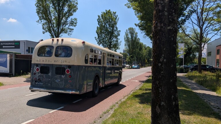 Klassieke bus met opschrift "n.h.a.d.o nv bergen" rijdt over een weg omgeven door bomen en gebouwen.