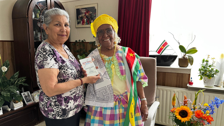Twee vrouwen poseren in een woonkamer, een met kleurrijke traditionele kleding en een Surinaamse vlag. Ze houden samen een certificaat vast.