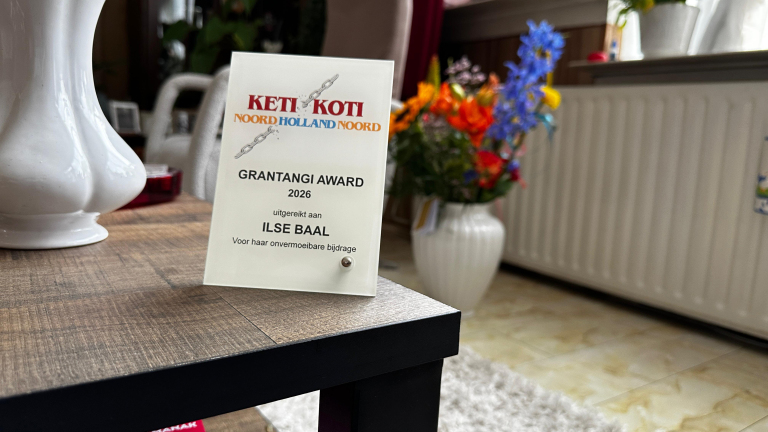 Een certificaat voor de Keti Koti Grantangi Award 2026 uitgereikt aan Ilse Baal, geplaatst op een houten tafel nabij een bloemenvaas.