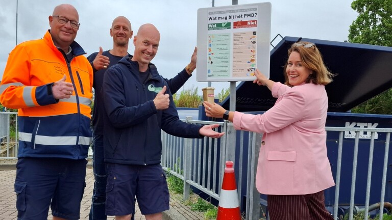 Vier mensen poseren voor een PMD-informatiebord bij een afvalcontainer, waarbij ze goedkeurend gebaren en lachen.