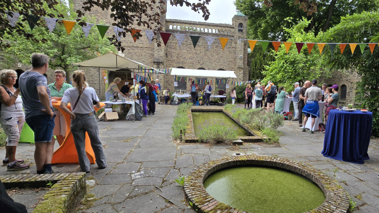 Mensen praten en winkelen bij een buitenmarkt met kleurrijke vlaggetjes, tafels en een tuin met vijver omgeven door historische gebouwen.