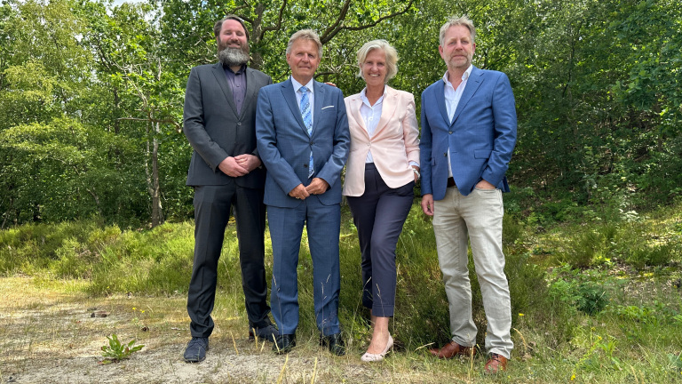 Vier mensen in zakenkleding poseren buiten op een grasveld met bomen op de achtergrond.