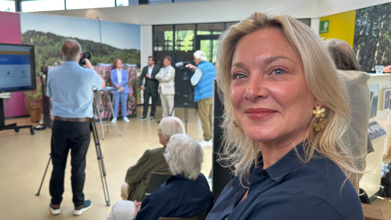 Vrouw glimlachend naar de camera in een zaal met een presentatie op de achtergrond.